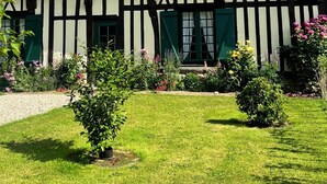 Una casa tradicional con entramado de madera y techo inclinado, dos ventanas dormilonas y un jardín bien cuidado con diversas plantas y flores.