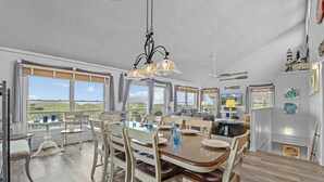 A dining area with a large table, chairs, and a chandelier. There are windows with a view of the outdoors. - Oceanfront Home in Waves with Pool, Hot Tub, and Game Room (Waves)