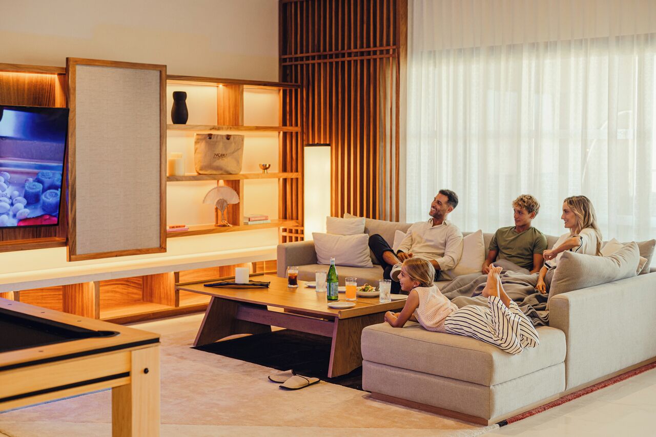 Un salon moderne avec un canapé, une table basse et un téléviseur. On y trouve des étagères en bois ornées d’objets décoratifs et une grande fenêtre avec des rideaux légers.