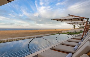 Zona junto a la piscina con sillones de descanso y sombrillas, con vistas a una playa.