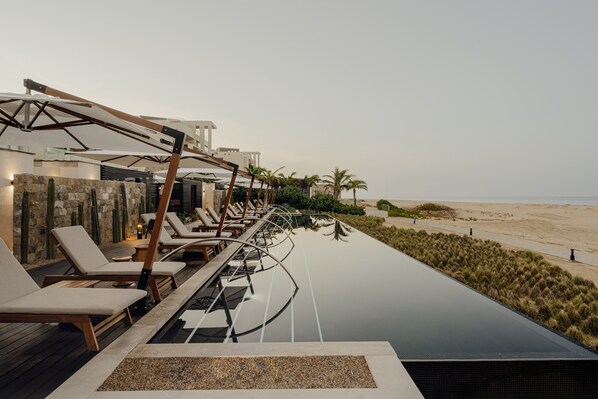 Una piscina infinita con sillones de descanso, vista a la playa y un paisaje desértico.
