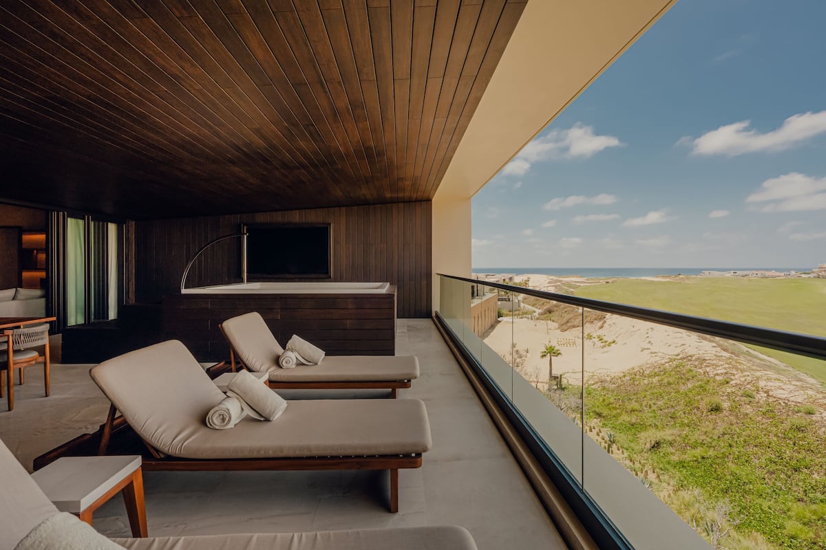 A balcony with two lounge chairs, a small table, and a TV. The balcony overlooks a beach and ocean.
