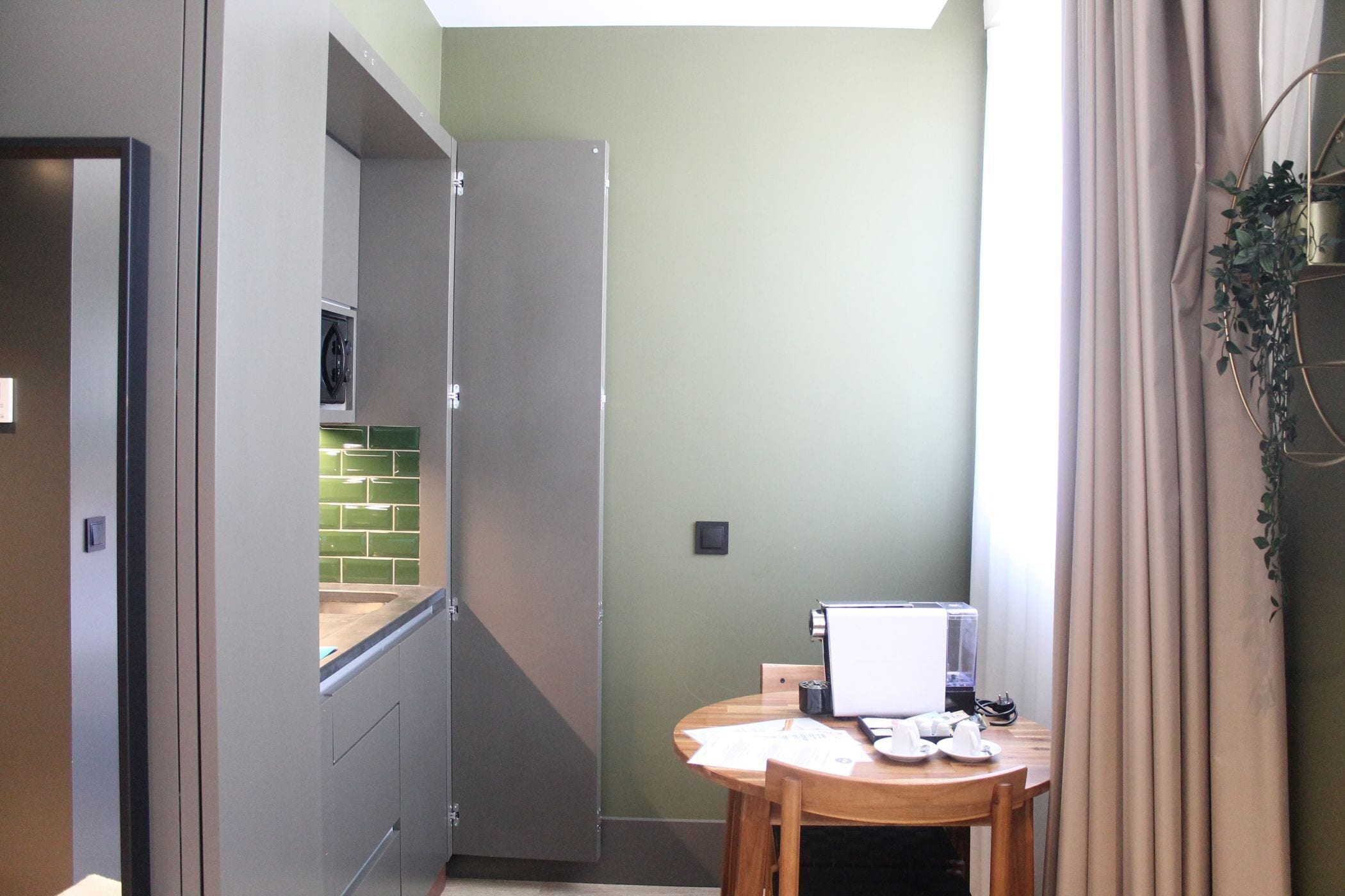 A compact kitchen with a refrigerator, a microwave, and a small window. There is a dining table with chairs and a laptop.