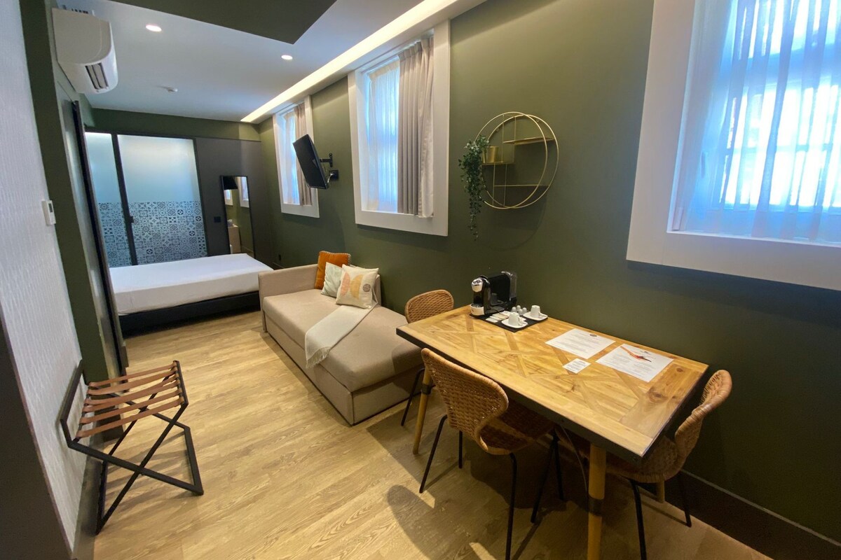 A compact room with a bed, a sofa, a dining table with chairs, a small kitchen area, and a window with blinds.