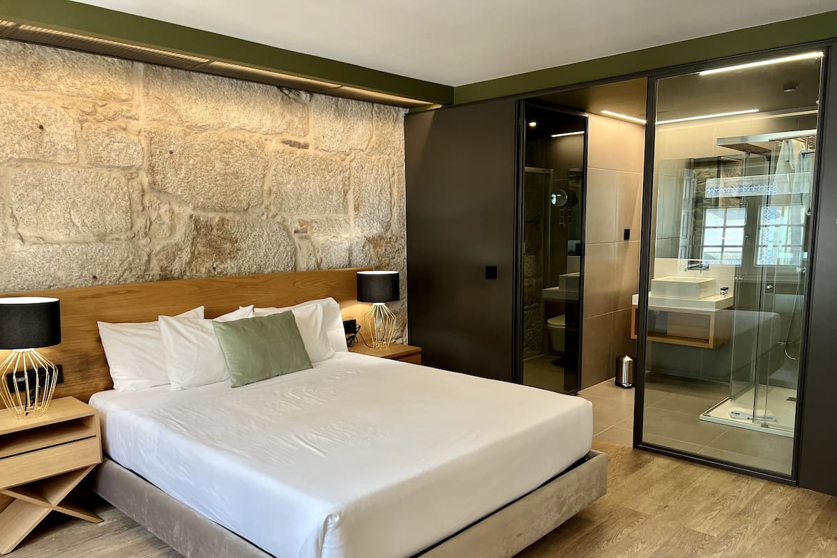 A modern hotel room with a large bed, a stone accent wall, a glass-enclosed bathroom, and a wooden floor.