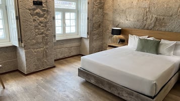 Un dormitorio con pared de piedra, una cama con ropa de cama blanca, una mesita de noche con una lámpara y una ventana con vistas.