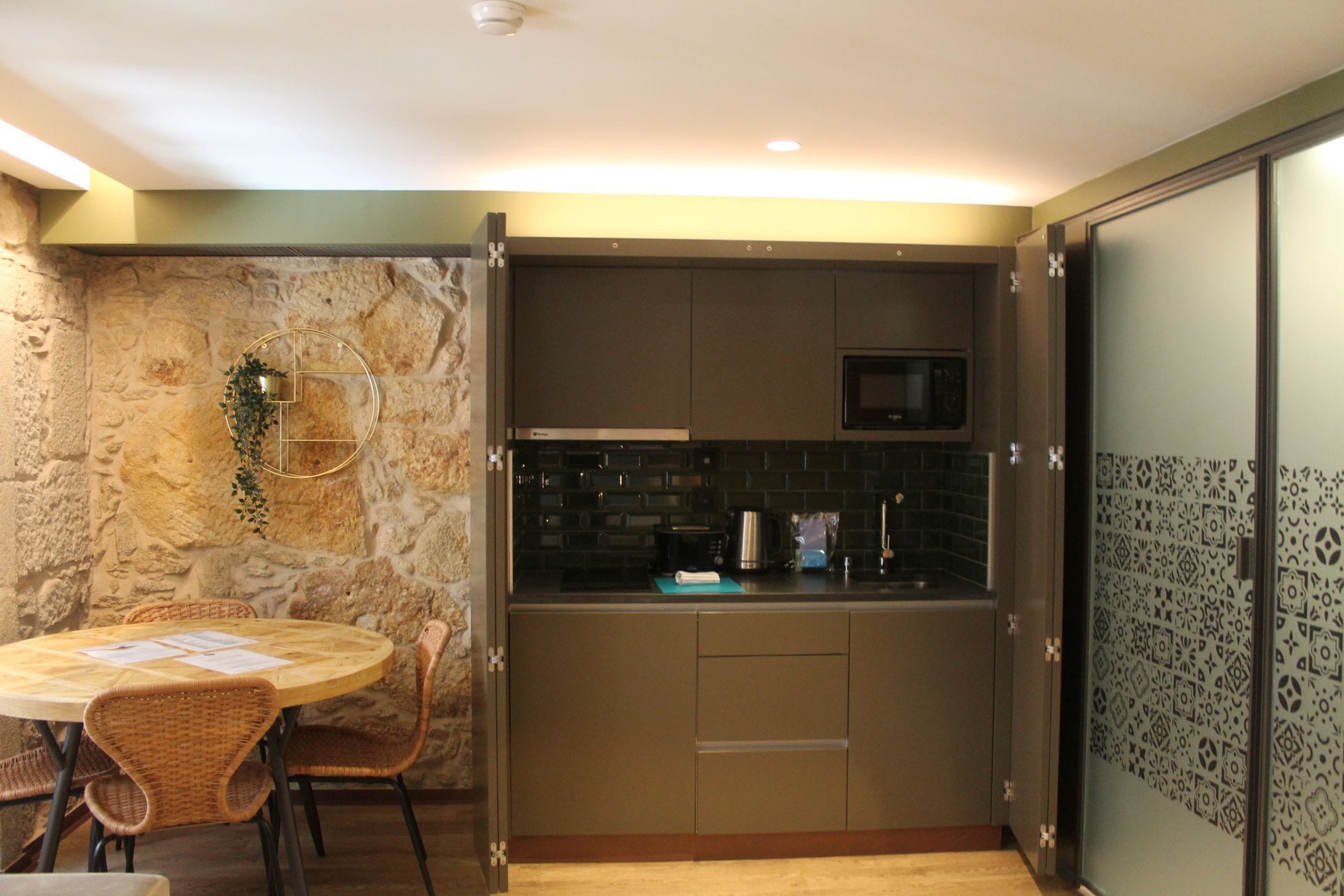 Una cocina compacta con pared de piedra, microondas y un pequeño comedor con sillas de mimbre.