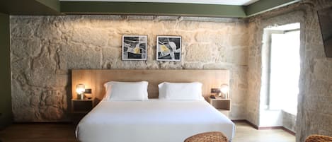 A hotel room with a stone wall, a large bed with white linens, two bedside tables with lamps, and a view of the outdoors.