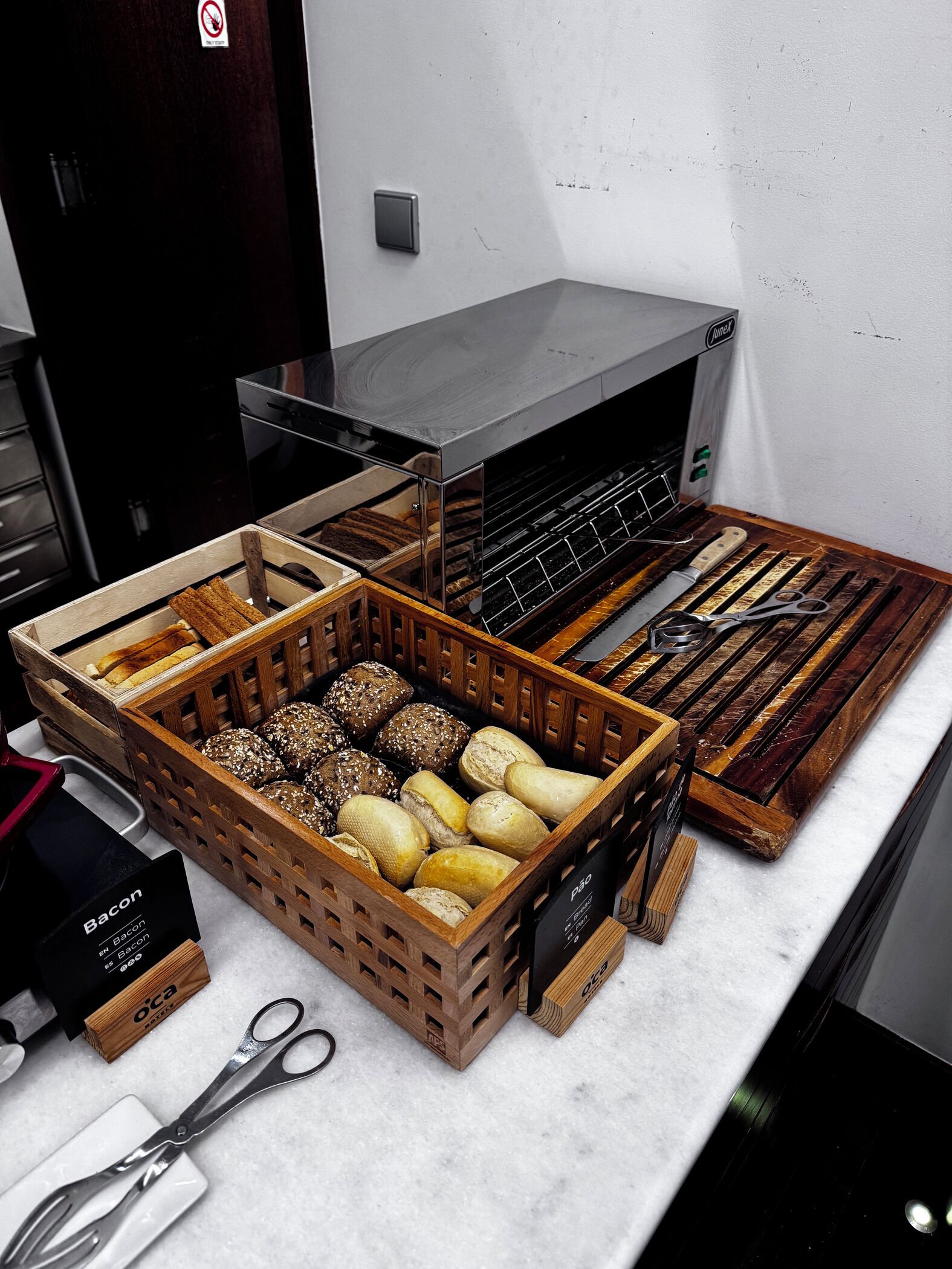 A kitchen counter with various types of bread and pastries in wooden crates, a box labeled "Bacon," and a pair of scissors.