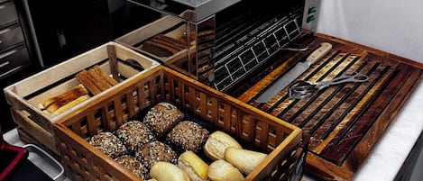 A kitchen counter with various types of bread and pastries in wooden crates, a box labeled "Bacon," and a pair of scissors.