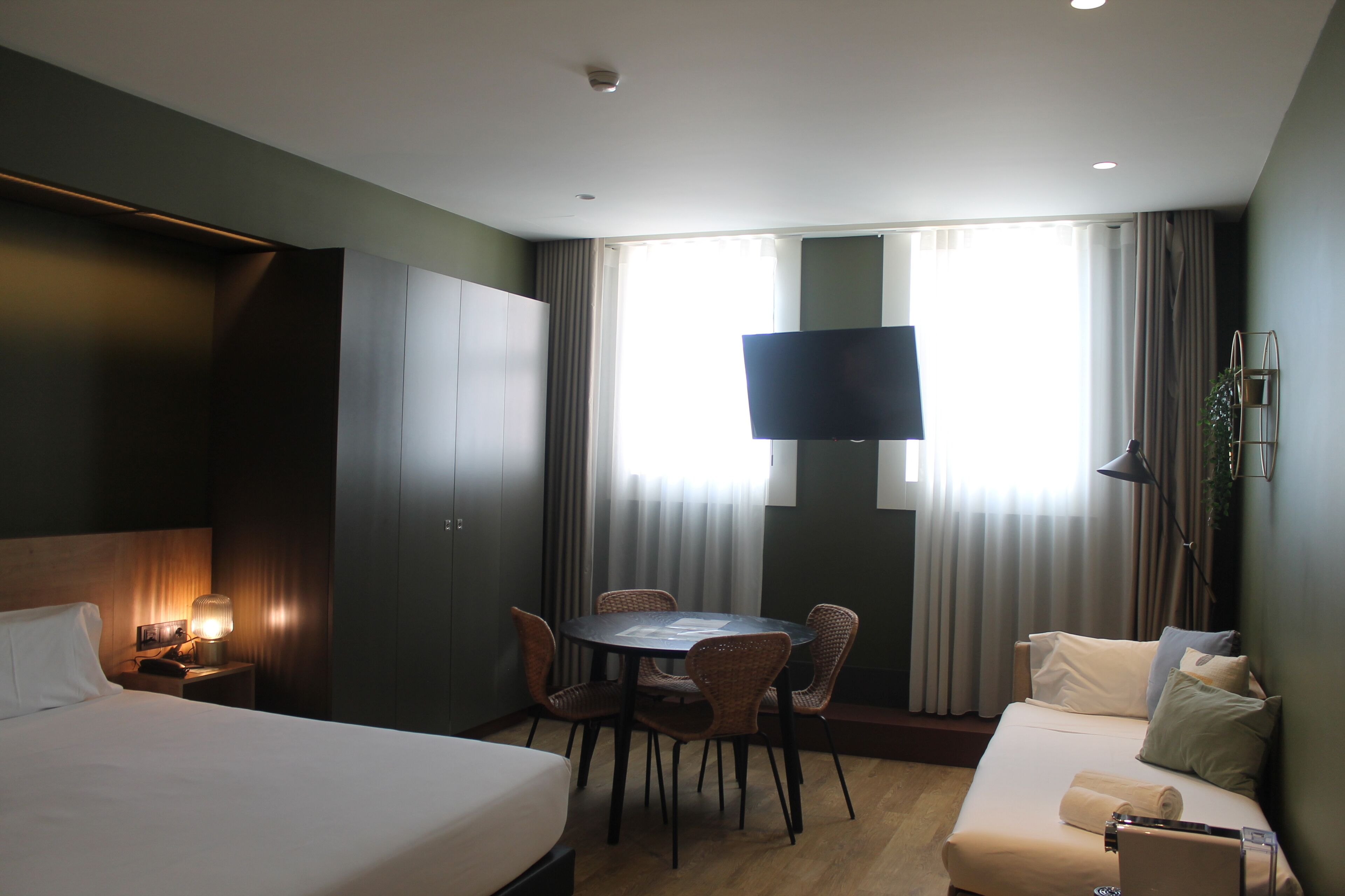 Een hotelkamer met een bed, een bank, een eettafel met stoelen, een aan de muur bevestigde televisie en een raam met gordijnen.