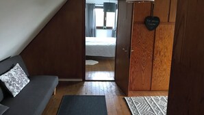 Tunnelmallinen ullakkohuone, jossa on kalteva katto, puinen vaatekaappi, harmaa sohva ja kuviollinen matto.