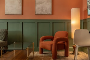 Um quarto com painel verde, duas cadeiras laranja, uma cadeira branca, uma mesa de vidro e um abajur.