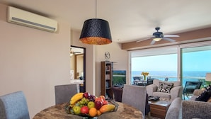 A dining area with a fruit bowl on a round table, surrounded by chairs. There is a ceiling fan and a view of the ocean through large windows. - Modern- Renovated- Breathtaking 6th Floor- 3 Bedrooms Villa Magna (Nuevo Vallarta)