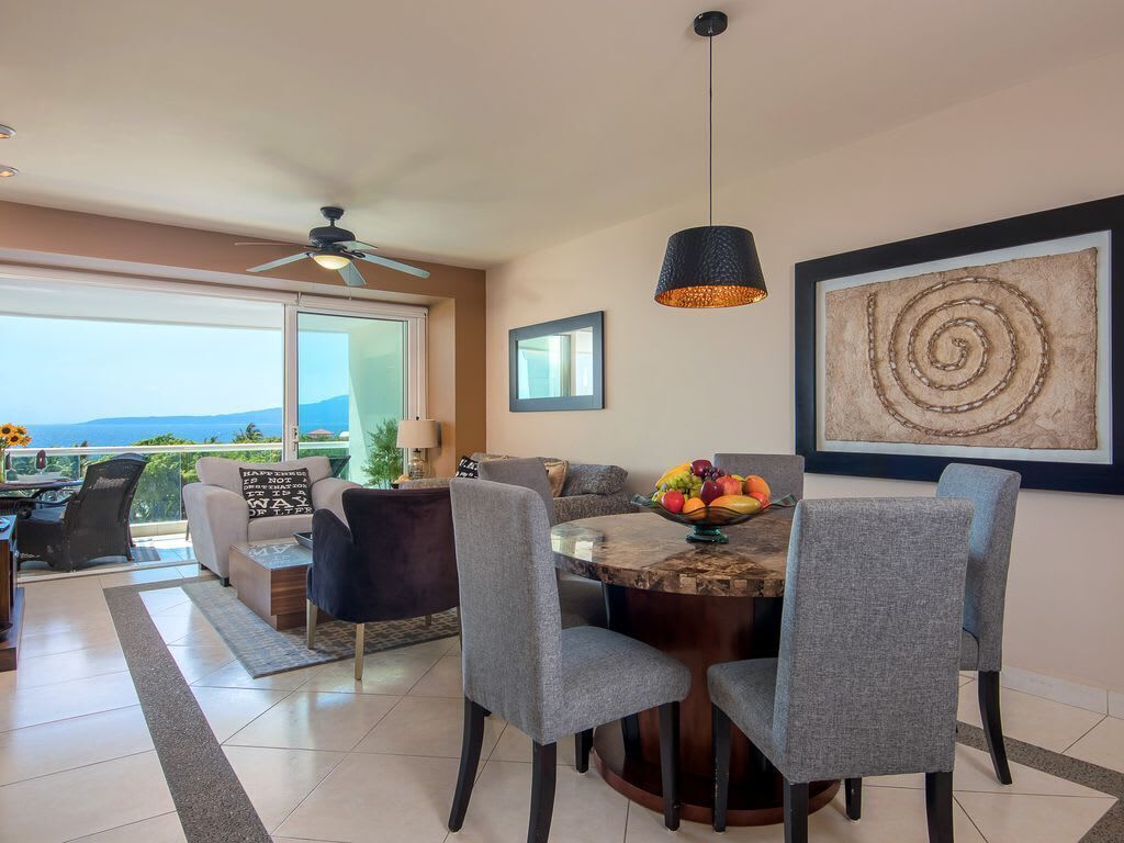 A dining area with a round table, grey chairs, a fruit bowl, and a pendant light.