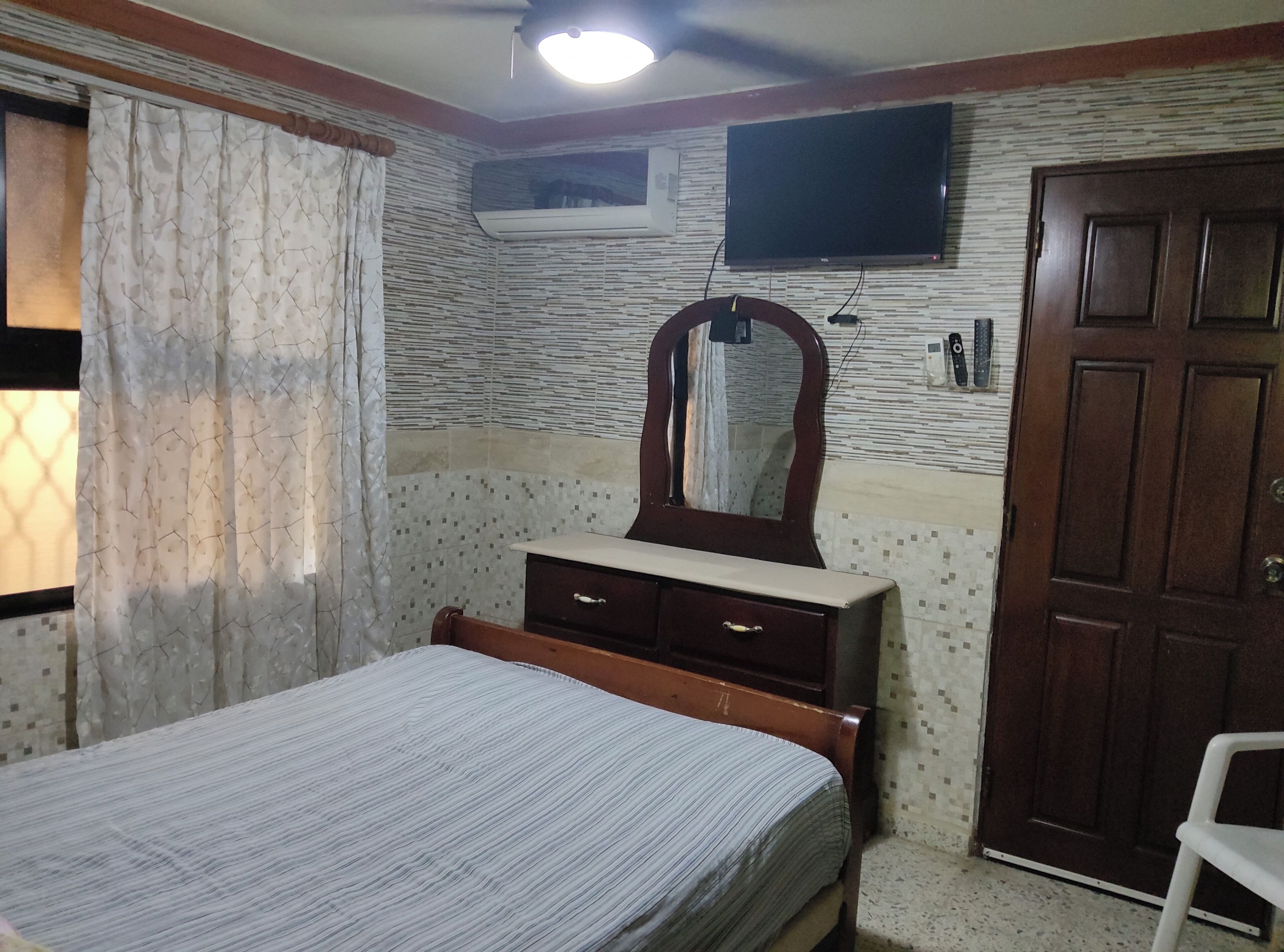 A bedroom with a bed, a dresser, a mirror, an air conditioning unit, a television, and a door.
