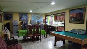A room with a pool table, dining area with chairs and a table, kitchen area with cabinets, and a refrigerator.