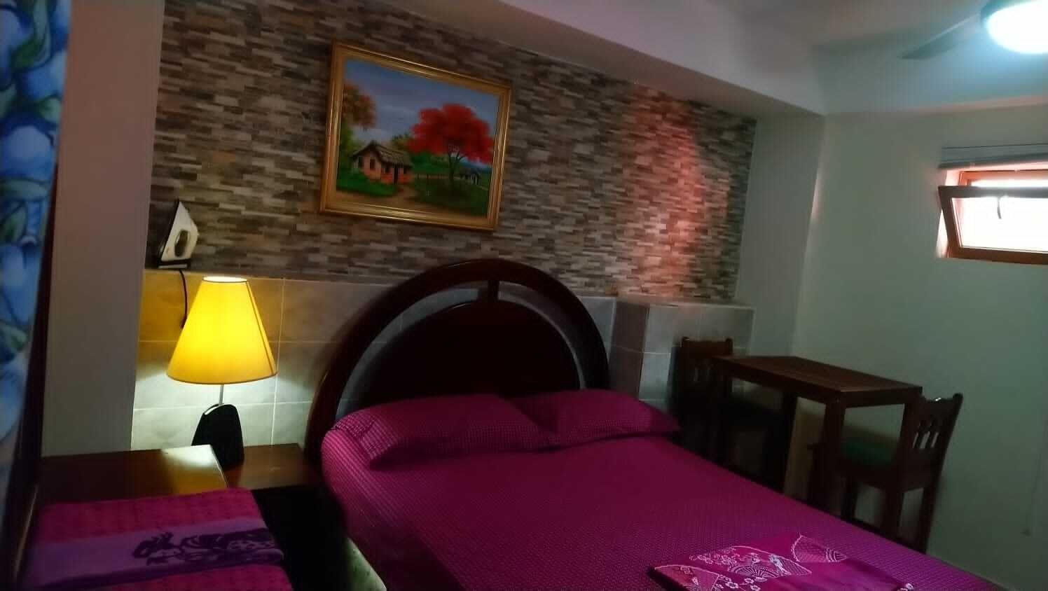 A bedroom with a bed, a nightstand, a lamp, a painting on the wall, a window, and a wooden table.
