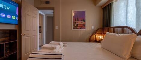 A hotel room with a bed, a TV displaying the Apple TV interface, a door leading to a hallway, and a framed picture on the wall.