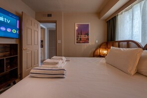 A hotel room with a bed, a TV displaying the Apple TV interface, a door leading to a hallway, and a framed picture on the wall.