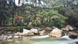 A natural setting with a stream, rocks, and trees. There are structures with red roofs in the background. - Athaka'i (Concepción)