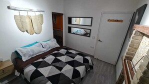 A bedroom with a bed, a wooden headboard, a wall-mounted shelf with decorative items, a door, and a brick wall. - Athaka'i (Concepción)