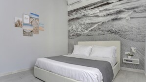 A modern bedroom with a large wall mural of ocean waves, a neatly made bed with white bedding, and a small bedside table with a potted orchid. - Voyager House (Rome)