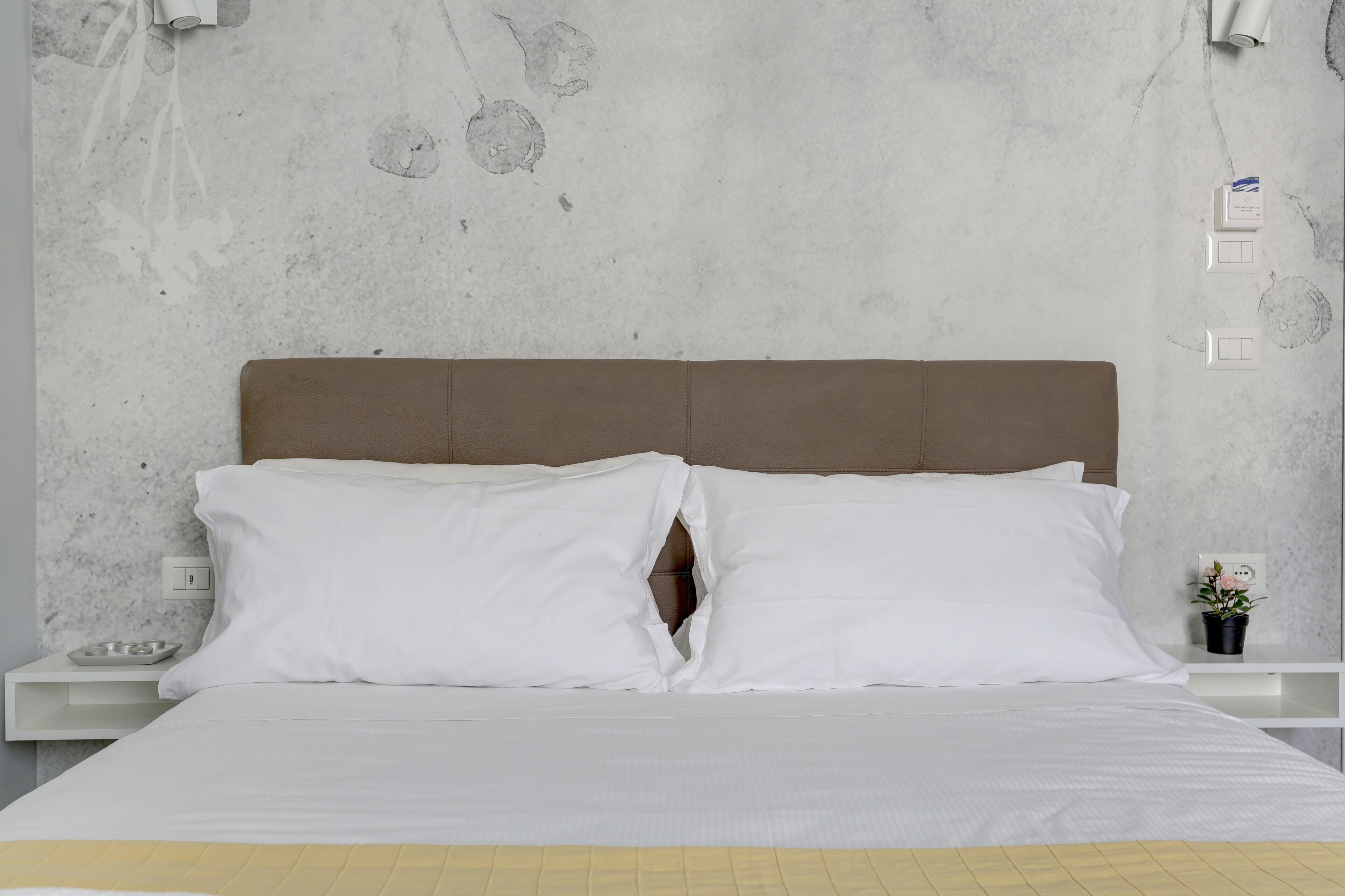 Una cama con ropa de cama blanca y cabecera marrón, una mesita de noche pequeña con lámpara, y una pared con textura y un enchufe.