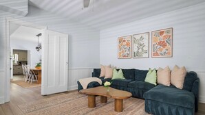 Ein Wohnzimmer mit einem dunkelgrünen Sofa, einem Holztisch und einem gemusterten Teppich. An der Wand hängen drei gerahmte Blumenbilder.