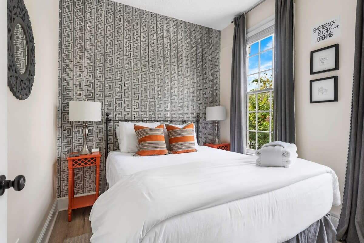 Un lit bien fait, avec du linge de lit blanc et des coussins à rayures, une table de chevet avec une lampe, un mur orné d’un motif géométrique et une fenêtre avec des rideaux.