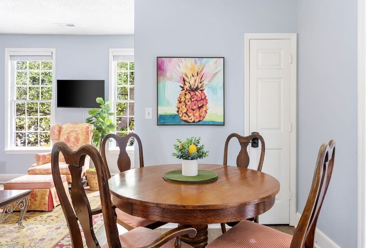 A dining area with a round wooden table, six wooden chairs, and a painting of a pineapple on the wall.