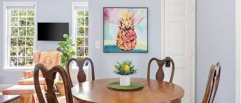 A dining area with a round wooden table, six wooden chairs, and a painting of a pineapple on the wall.