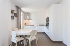 Et moderne køkken med hvide skabe, en indbygget ovn og spiseplads med et rundt bord og hvide stole.