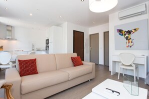 A modern living room with a beige sofa, red and white patterned pillows, a glass coffee table, and a white desk with a chair. - Premium Design Renovated 2 BR 2 bath Croisette 200 m Olam Properties (Cannes)