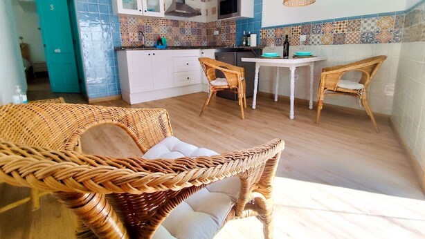 Una cocina con armarios blancos, microondas y estufa. Hay sillas de mimbre y una mesa.