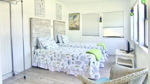 Un dormitorio con dos camas, un ventilador de techo, una silla de mimbre y una ventana con persianas.