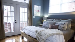 Quarto com cama de madeira, mesa de cabeceira, abajur, janela com persianas e porta com painéis de vidro.
