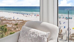 A room with a large window overlooking a beach, a white sofa with a starfish pillow, and a view of the ocean and beach activities. - Gulf Front | Fully Renovated | Pool and Hot Tub (Destin)