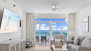 A spacious living room with a large window, a ceiling fan, a flat-screen TV, a sofa, a coffee table, and a view of the ocean. - Gulf Front | Fully Renovated | Pool and Hot Tub (Destin)