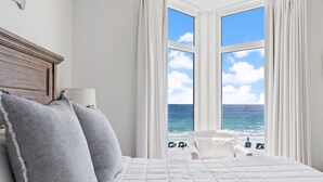A bedroom with a large window overlooking the beach, a bed with white bedding, and a chair by the window. - Gulf Front | Fully Renovated | Pool and Hot Tub (Destin)