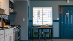 Een kleine keuken met een magnetron, fornuis en kastjes. Er is een raam met zonwering, een tafel met stoelen en een deur met een bordje.