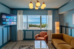 Habitación de hotel con un ventanal grande, un televisor de pantalla plana mostrando servicios de streaming, un sofá, una silla y una alfombra estampada.