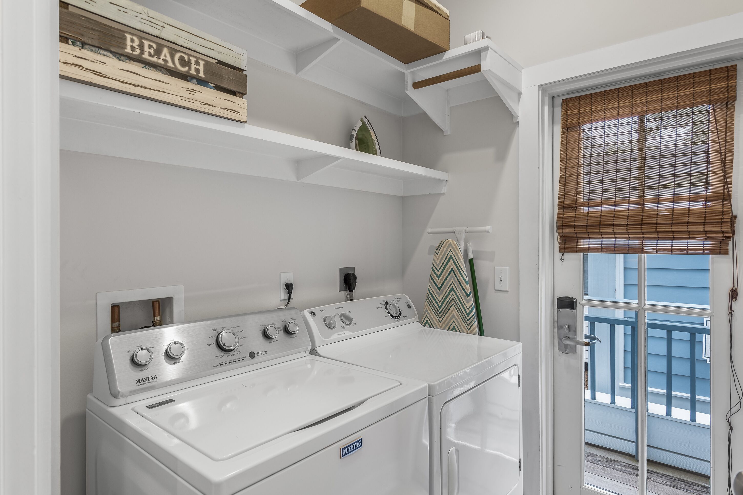 A laundry area with a Maytag washer and dryer, a wooden shelf with a 'BEACH' sign, and a window with blinds.
