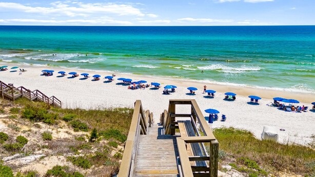 A beach with a wooden boardwalk leading to the shore, lined with blue umbrellas and beach chairs.