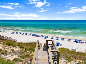 A beach with a wooden boardwalk leading to the shore, lined with blue umbrellas and beach chairs. - Magnolia Cottages by the Sea by PHG (Panama City Beach)