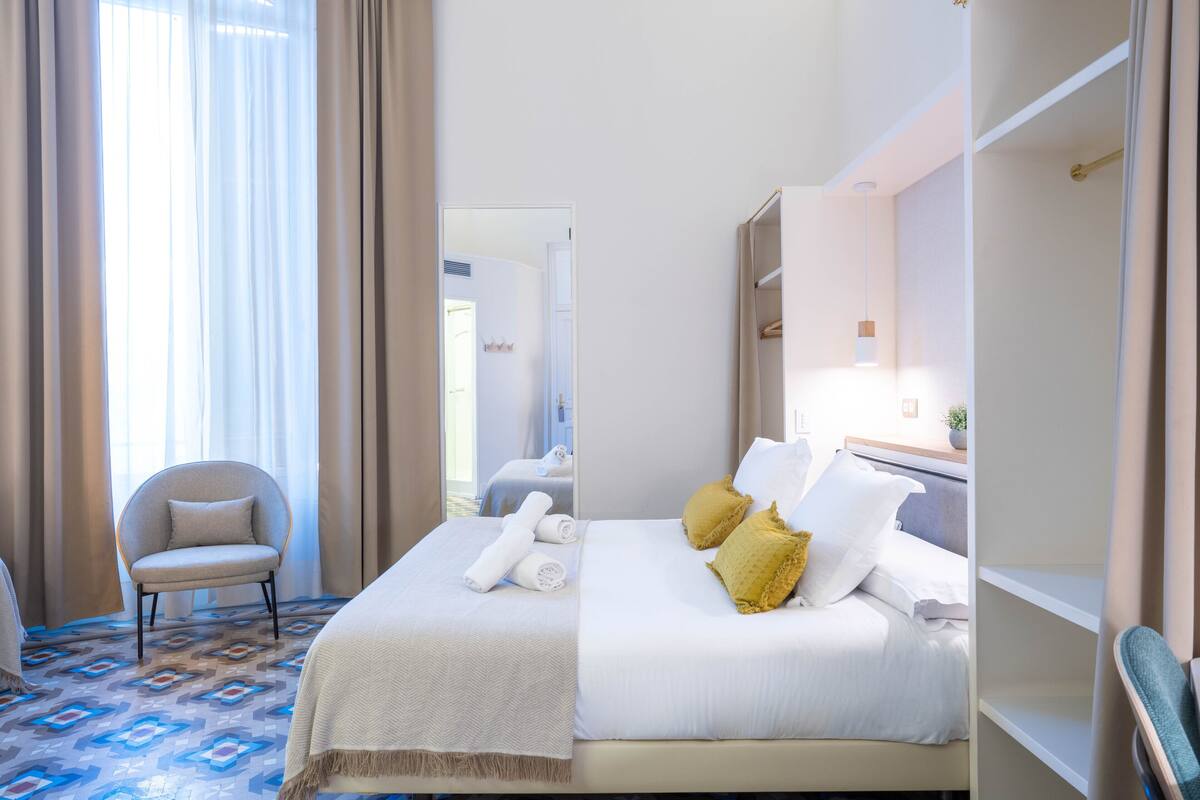 A hotel room with a bed, a chair, a mirror, and a window with curtains.