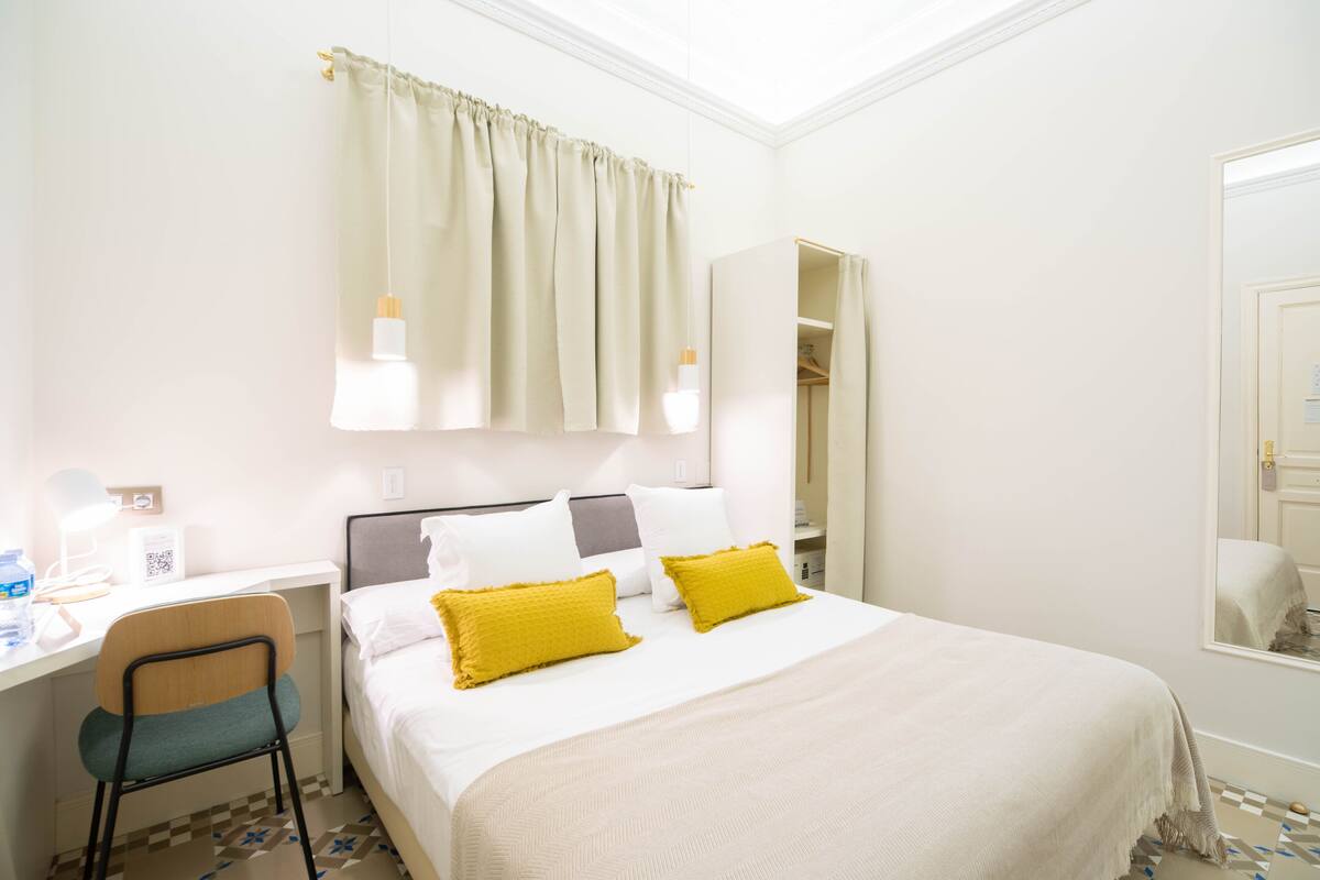 A hotel room with a bed, a desk, a chair, a mirror, and a window with curtains.