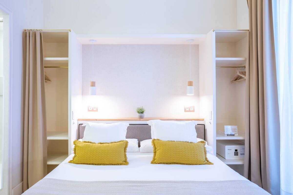 A hotel room with a bed, two bedside tables, a nightstand, a wall-mounted lamp, a small potted plant, and a closet with hanging clothes.
