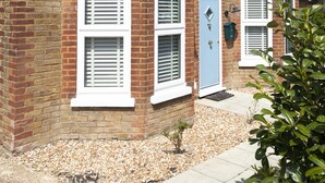A two-story brick house with white window frames and shutters, a blue door, and a small garden area with gravel and a potted plant. - Sunbeam and Sundance Cosy House (whitstable)