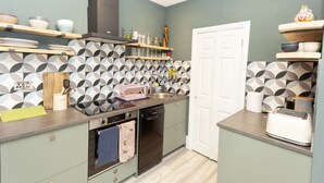 A compact kitchen with a dishwasher, oven, and a toaster. There are shelves with dishes and a variety of kitchen utensils. - Sundance Cosy Getaway (whitstable)